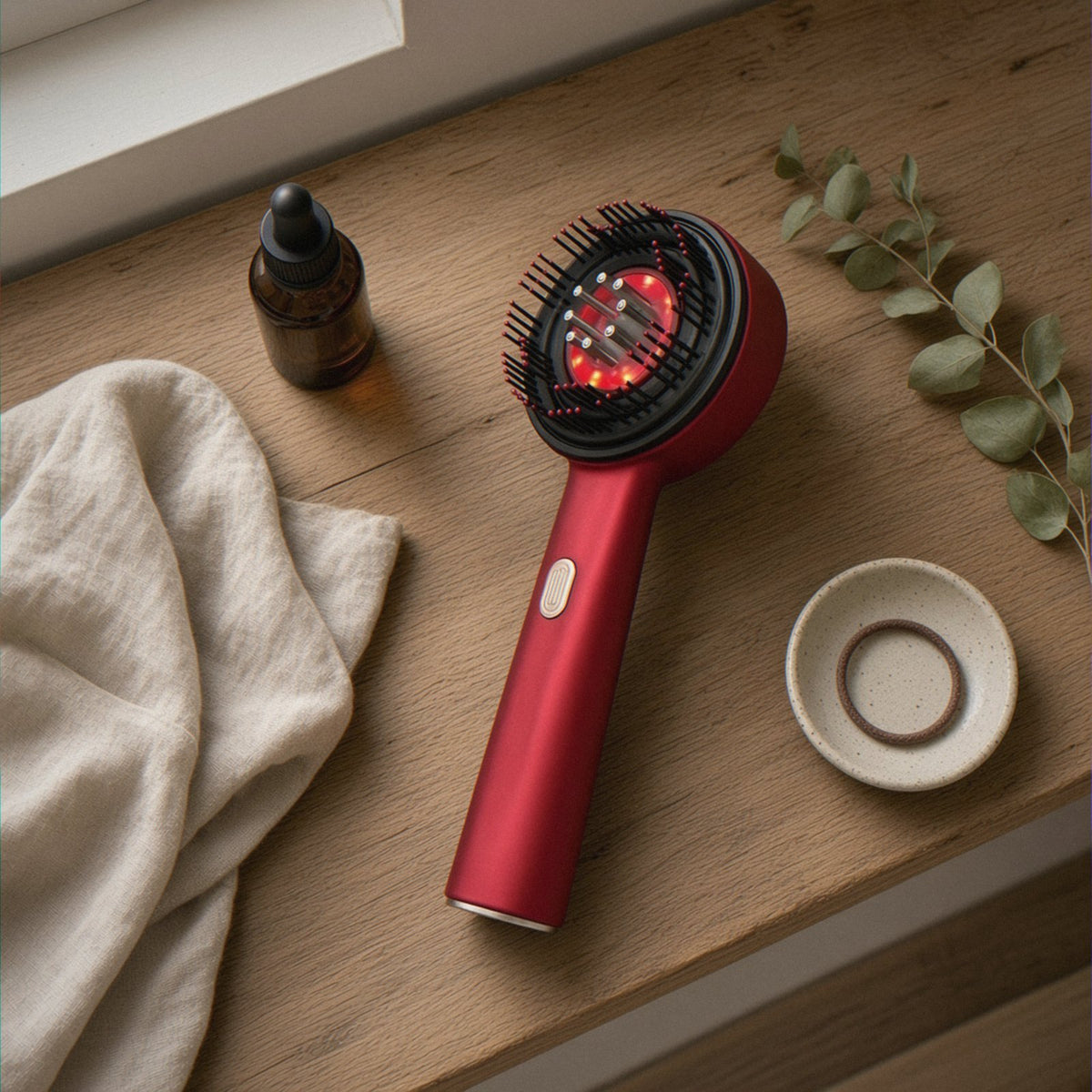 Flat lay of the 3-in-1 Red Light Scalp Massaging Brush on a wooden shelf with an amber oil bottle, linen and eucalyptus